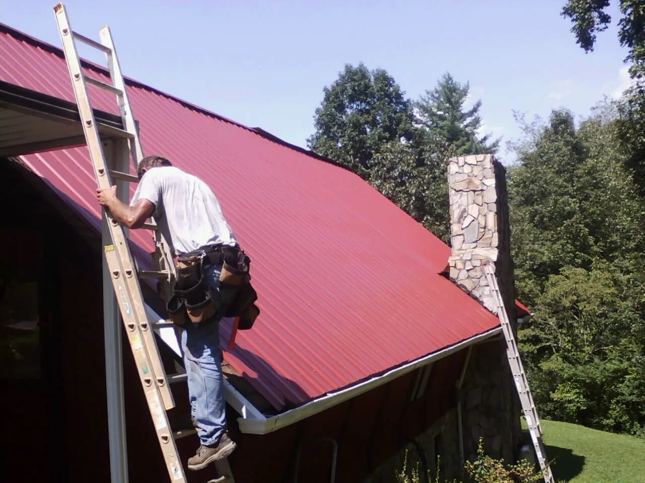 Roofer on a ladder beside a red metal roof and stone chimney — Soffit & Fascia Repair by Roofing Brothers in Otis
